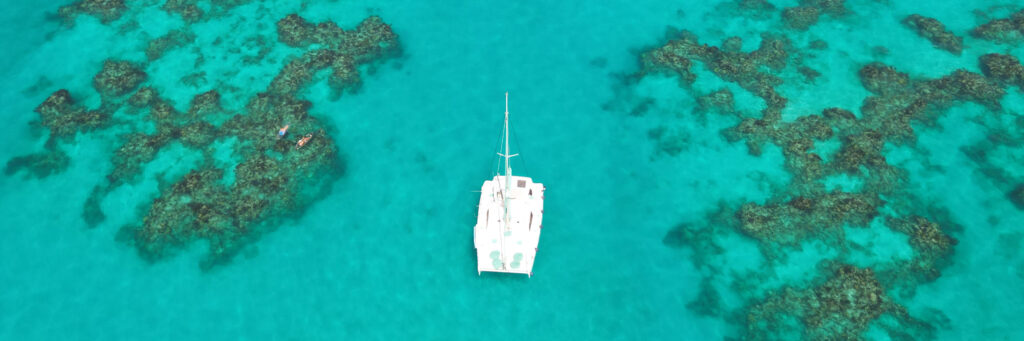 A white catamaran floats on clear turquoise water, surrounded by patches of coral reef visible beneath the surface. The scene is viewed from above, capturing the boat and the vibrant, shallow sea.