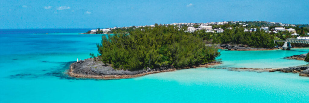 A small, tree-covered island sits in bright turquoise water, with a bridge connecting it to the mainland. White houses dot the distant green shoreline under a clear blue sky.