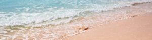 A gentle wave rolls onto a sandy beach with clear turquoise water meeting soft pink sand under a bright, sunny sky. The foamy edge of the wave creates a serene and tranquil scene typical of a tropical shoreline.