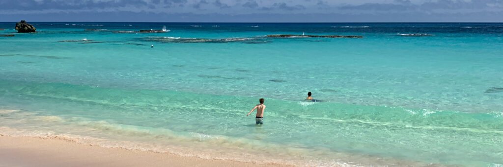 Two people wade in clear turquoise ocean water near a sandy beach under a cloudy sky. Small waves gently roll toward the shore, and rocks are visible in the distance.