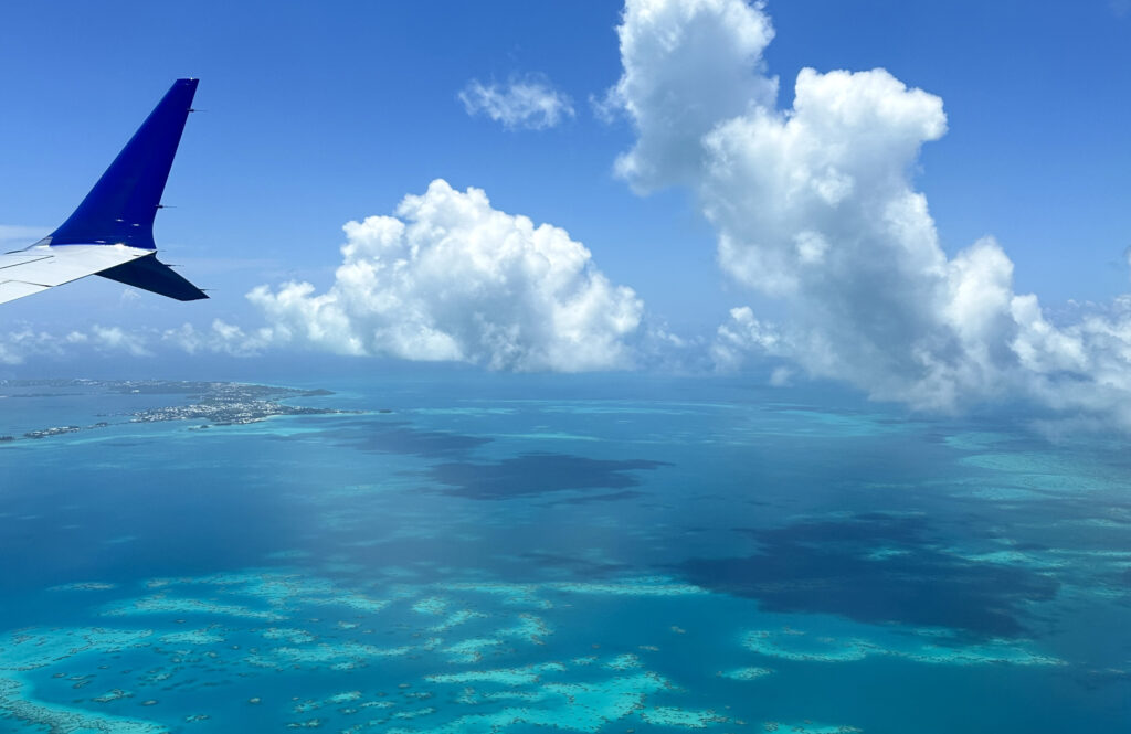 Aerial view from an airplane window showing a wingtip, bright blue sky with large clouds, and turquoise ocean water below with a distant coastline—perfect inspiration as you plan what to take on a cruise to Bermuda.