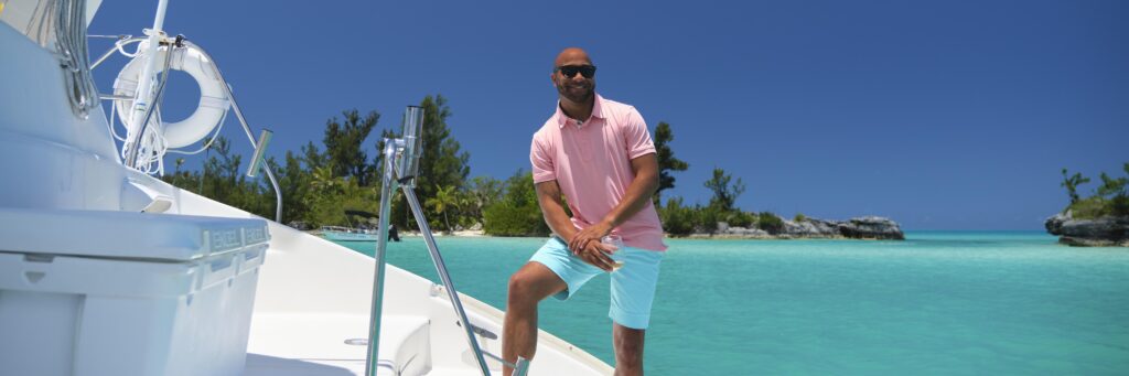 A man in a pink polo shirt and turquoise shorts stands smiling on a white boat, surrounded by clear turquoise water and lush greenery under a bright blue sky, ready to experience Bermuda Customs.