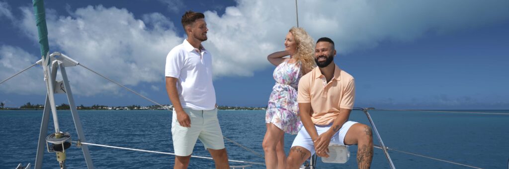 Three people, two men and one woman, are smiling and enjoying a sunny day on a sailboat with bright blue water and a partly cloudy sky—giving great inspiration for what to take on a cruise to Bermuda.