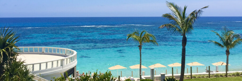 View of a tropical beach with turquoise water, a few palm trees, yellow beach umbrellas, and a white, curved terrace overlooking the ocean under a clear blue sky—truly capturing the relaxed spirit of Bermuda customs.