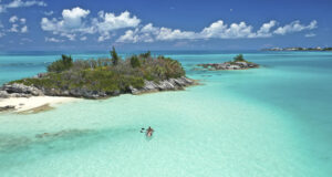 A person kayaks in clear turquoise water near a small, rocky island with lush greenery under a bright blue sky—an experience that highlights Bermuda must see attractions.