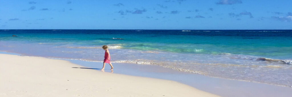 A child in a pink dress walks alone along a sunny, sandy beach beside clear blue ocean waves under a mostly clear sky—the scene captures the best time to visit Bermuda.