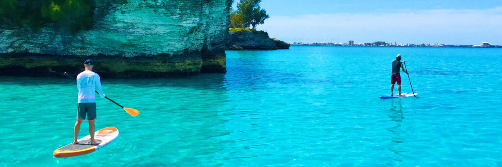 Two people paddleboarding on clear turquoise water near a rocky cliff with greenery; ideal conditions for Bermuda snorkeling, with a distant shoreline and buildings visible under a bright blue sky.