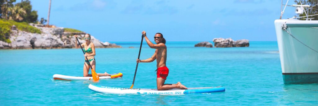 Two people paddleboarding on clear turquoise water—one kneeling and smiling at the camera, the other standing. A catamaran and rocky shoreline hint at perfect Bermuda snorkeling spots under a sunny sky.