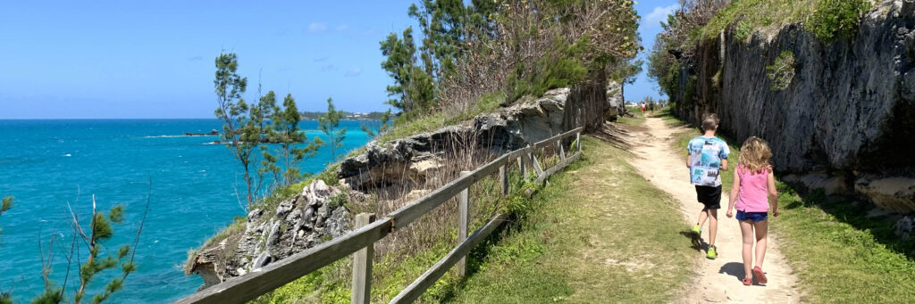 Two children walk along a dirt path by a wooden fence, with rocky cliffs, green trees, and the bright blue ocean of Bermuda Beaches under a clear sky on their left.