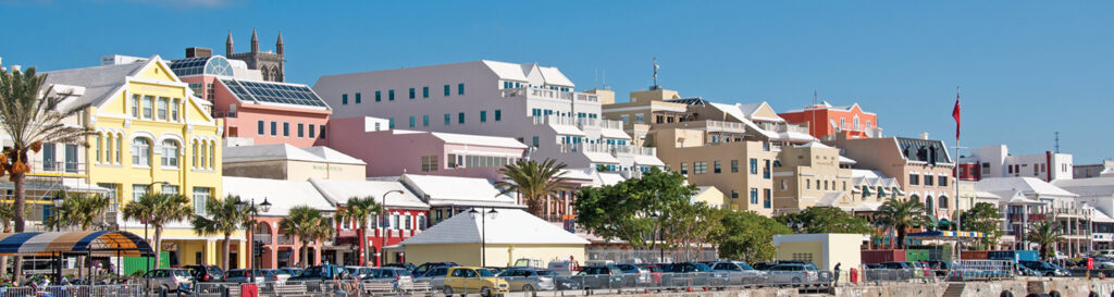 A vibrant harbor scene with colorful buildings along the waterfront under a clear blue sky. The diverse architecture and palm trees add charm, while cars line the street. This bustling area, home to some of the best places to eat in Bermuda, offers a perfect blend of scenery and flavor.