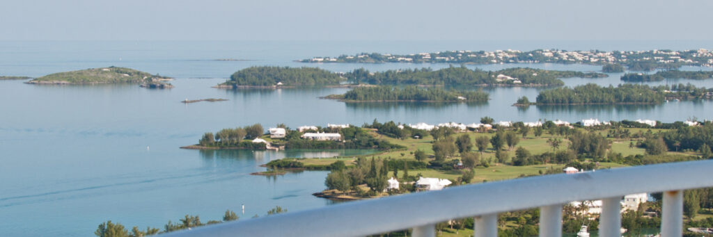A scenic view of calm blue water dotted with small green islands and white houses, seen from behind a white railing on a sunny day—one of Bermuda’s must see attractions.