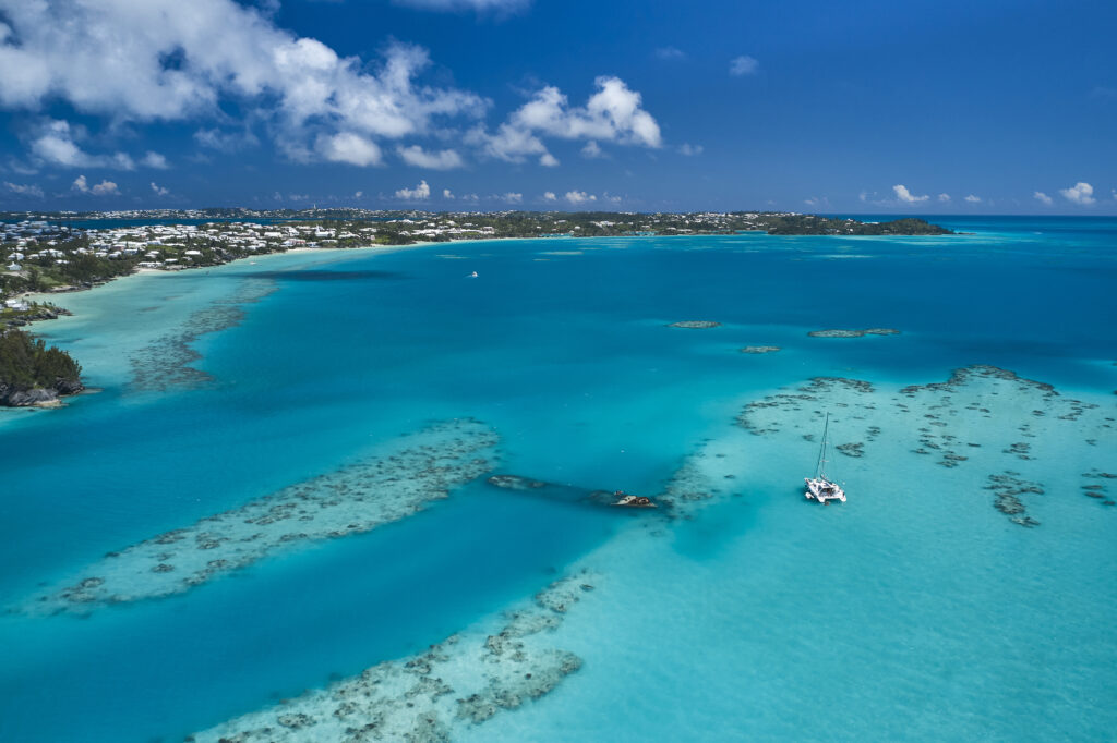 Aerial view of a turquoise ocean lagoon with a white catamaran, scattered coral reefs perfect for Bermuda Snorkeling, near a coastal town under a bright blue sky with scattered clouds.