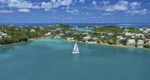 A catamaran floats on clear turquoise water, one of the Natural Wonders of Bermuda, near a lush, green coastline with scattered houses under a bright blue sky filled with fluffy clouds.