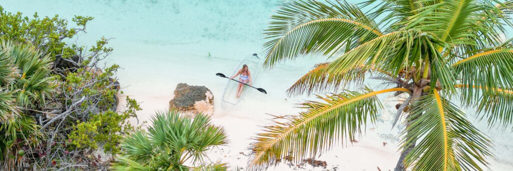 A woman sits in a clear kayak on turquoise water near a sandy shore, surrounded by palm trees and lush greenery—the perfect scene to experience during the best time to visit Bermuda.