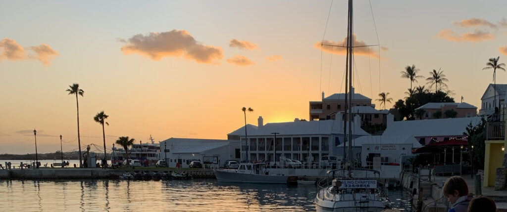 Sunset over a marina with boats docked by the waterfront, silhouetted palm trees, and pastel-colored buildings in the background under a partly cloudy sky—the perfect scene to capture during the best time to visit Bermuda.