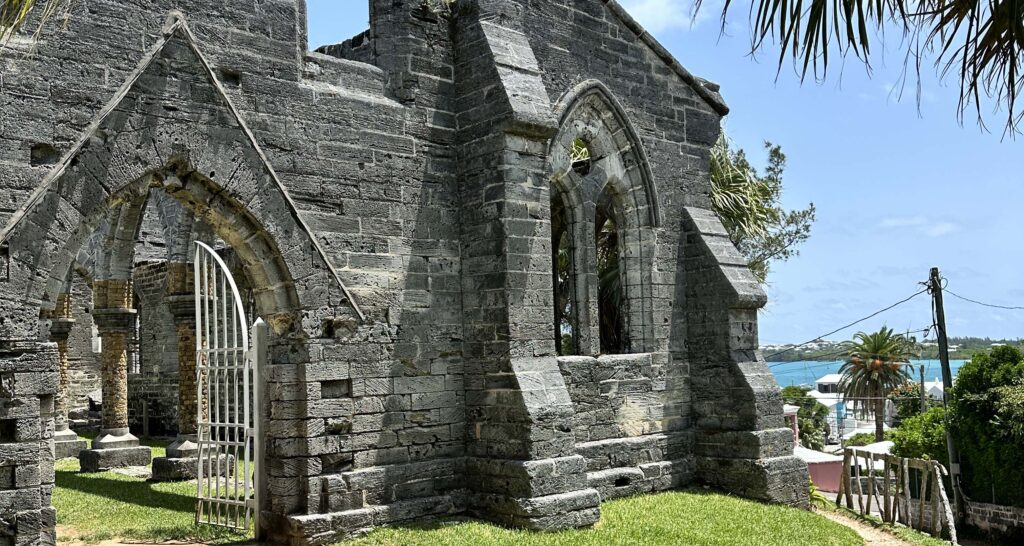 Stone ruins of a church with pointed arch windows and an open iron gate, set on green grass with palm trees and blue ocean in the background—one of the Natural Wonders of Bermuda under a sunny sky.
