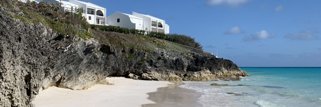 White buildings sit atop rocky cliffs overlooking Bermuda Beaches, where a sandy shore meets turquoise ocean under a clear blue sky, with gentle waves washing onto the coast.