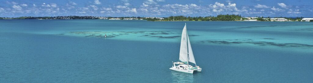 A white catamaran sailboat glides on clear turquoise water under a blue sky with scattered clouds, perfect for Bermuda snorkeling, with a distant shoreline and green trees visible in the background.