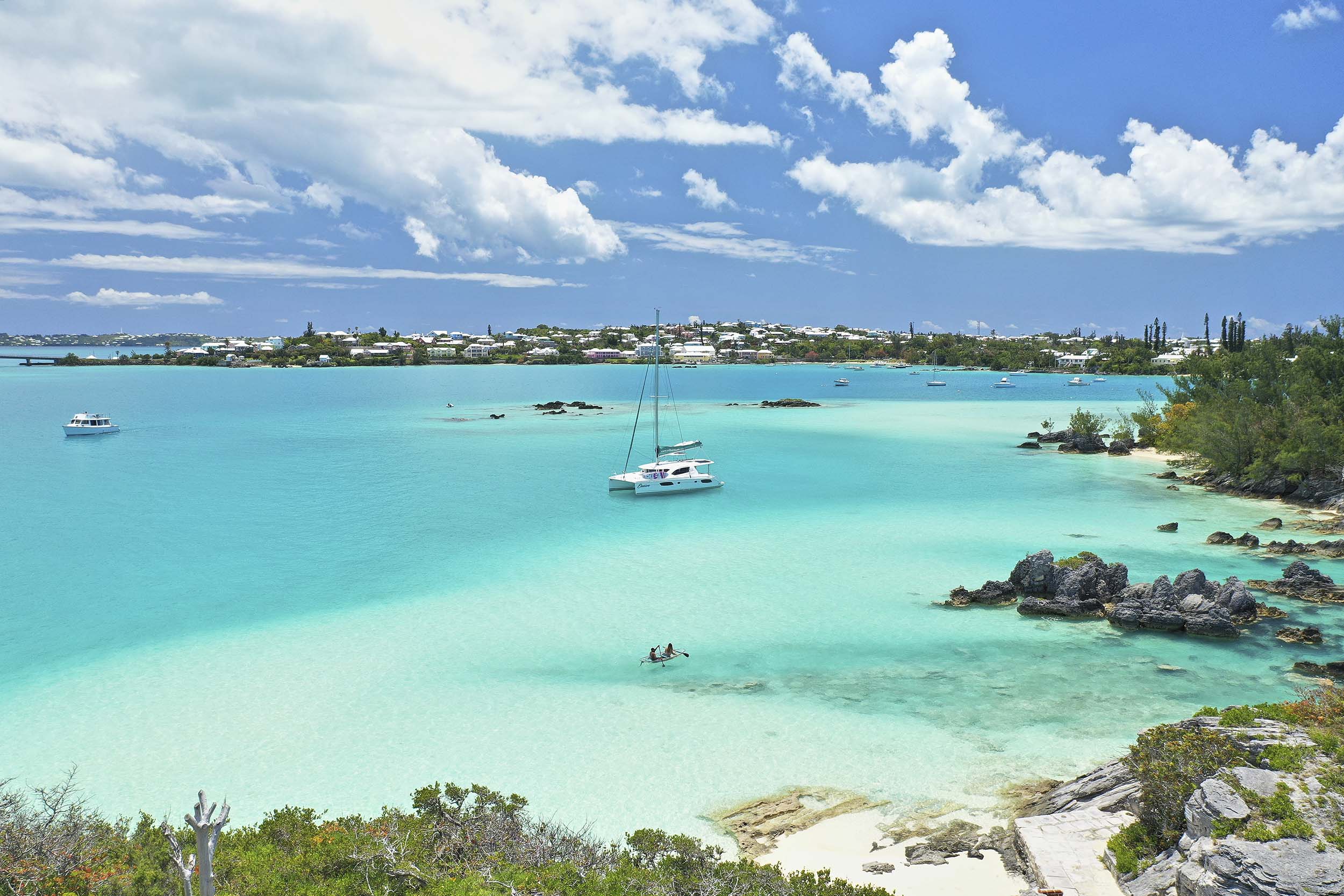 A turquoise bay with clear water, white sandy beaches, rocky outcrops, and boats anchored near the shore. Lush greenery surrounds the coastline under a blue sky with scattered clouds. Distant buildings dot the horizon.