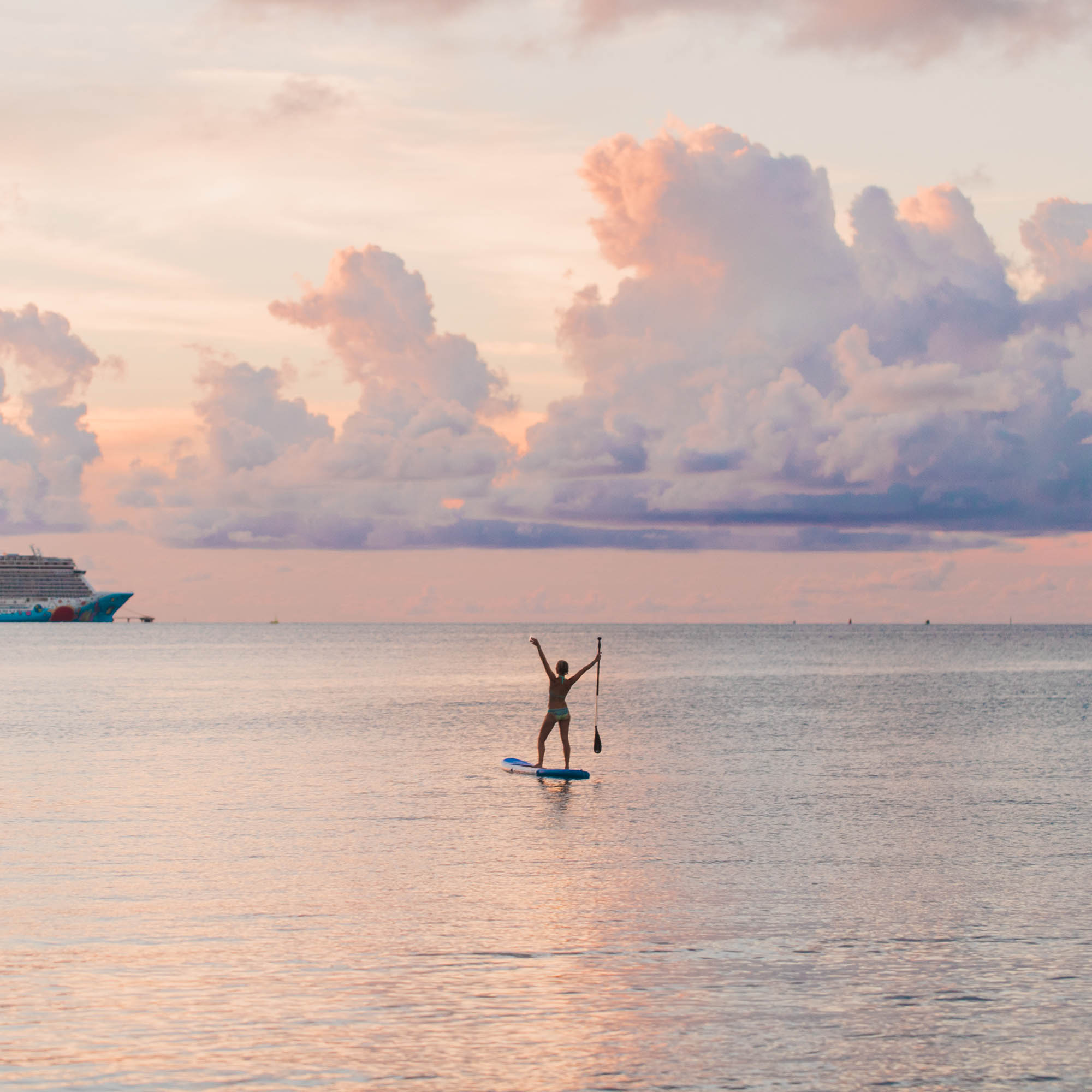 A person stands on a paddleboard in calm water at sunset, holding a paddle with arms raised. The sky is filled with colorful clouds, and a large ship is visible in the distance on the left.