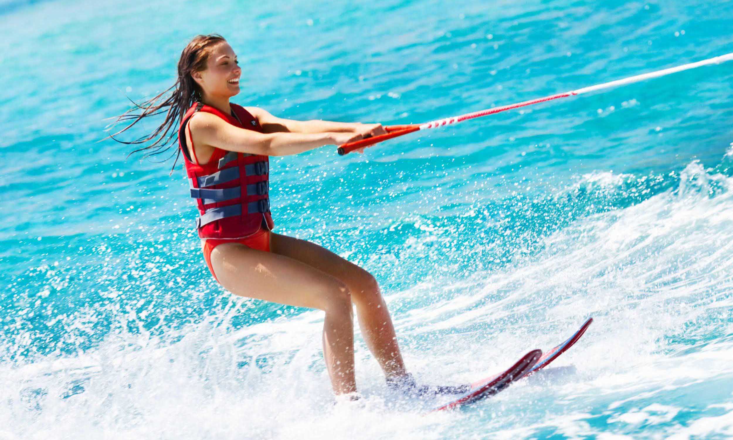 A young woman wearing a red life jacket is water skiing on bright blue water, holding onto a tow rope and smiling as she glides across the waves.