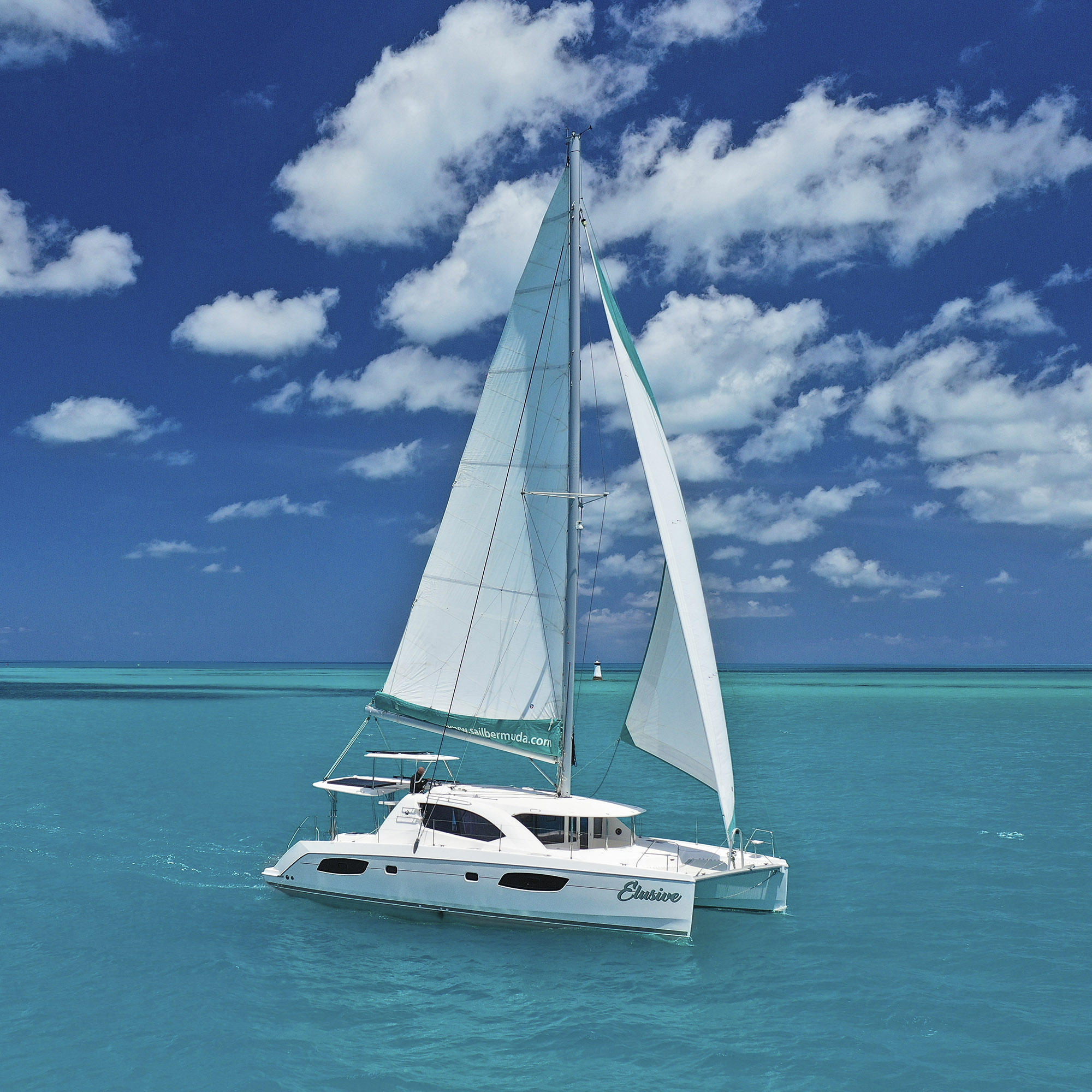 A white catamaran sailboat glides on clear turquoise water under a bright blue sky with scattered white clouds. The sails are fully up, and the sea appears calm and inviting.