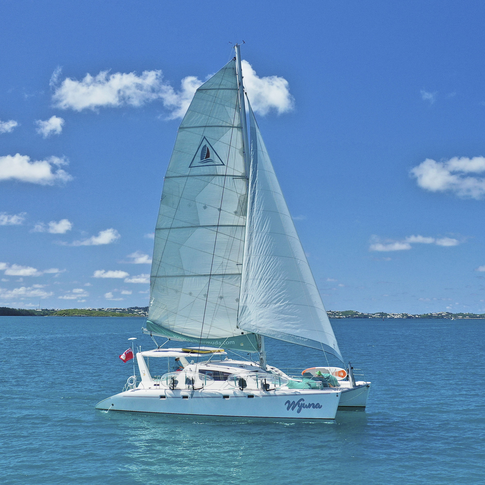 A white catamaran with the name Wyuna sails on clear blue water under a bright blue sky with scattered clouds. The coastline is visible in the background.