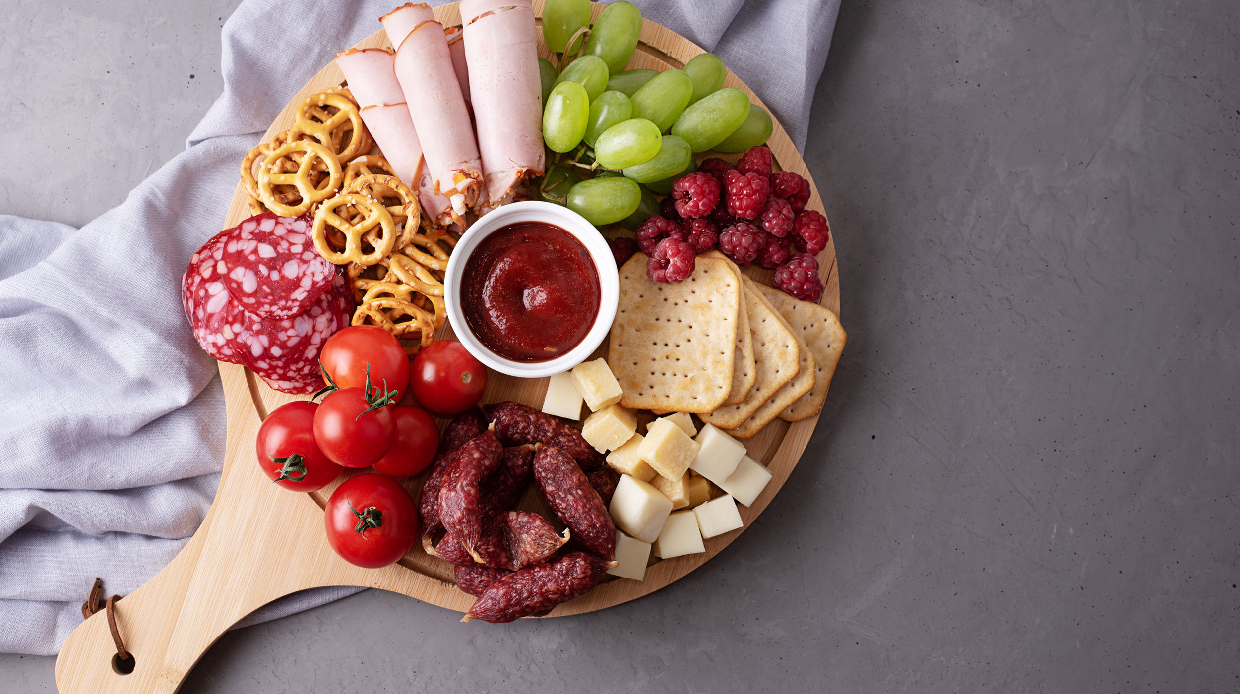 A wooden board with rolled deli turkey, salami, grape tomatoes, pretzels, cheese cubes, green grapes, raspberries, crackers, and a bowl of red dipping sauce, arranged on a gray surface with a gray cloth.