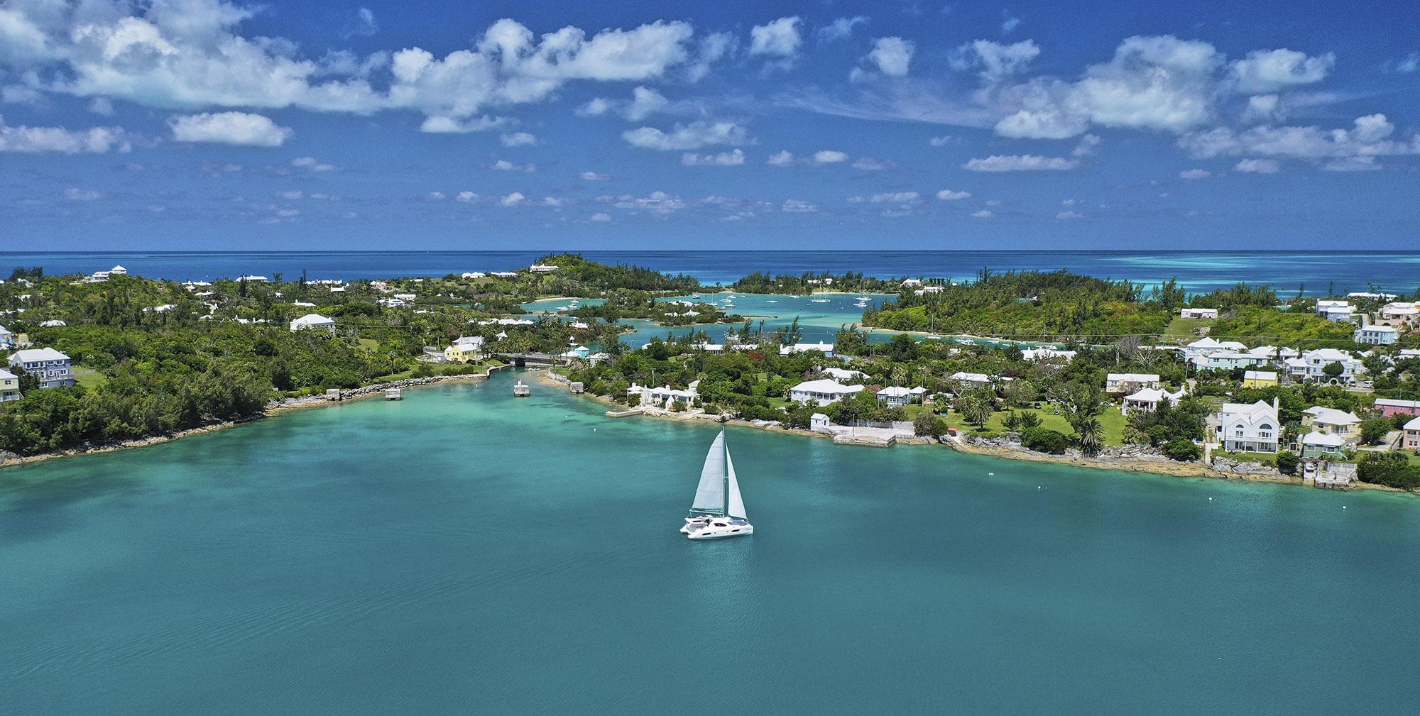 A catamaran with white sails gracefully glides across calm, turquoise waters under a bright sun, embodying the ideal weather in Bermuda for a perfect day on the sea. The scene captures the serene beauty of the ocean, making it an inviting sight for those looking to visit Bermuda during its warm summer months.