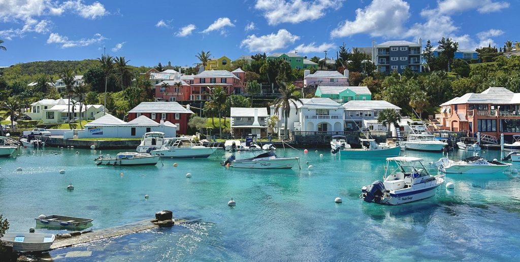 Colorful houses line a lush hillside above a clear blue harbor with boats anchored in calm water under a bright, partly cloudy sky. Palm trees and greenery are scattered throughout the scene.