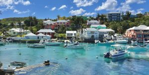 Colorful houses line a lush hillside above a clear blue harbor with boats anchored in calm water under a bright, partly cloudy sky. Palm trees and greenery are scattered throughout the scene.