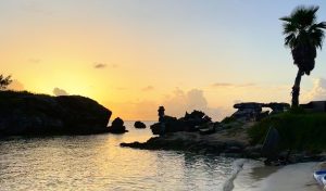 A tranquil beach scene at sunset, with calm water, rocky formations, and silhouettes of palm trees under a colorful sky fading from orange to blue.