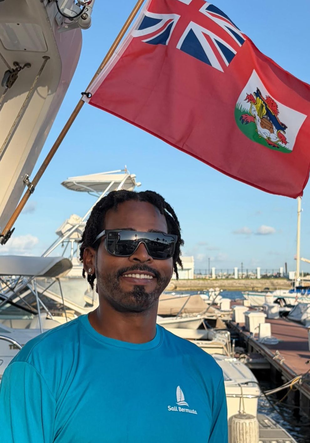 A man in a blue Sail Bermuda shirt stands on a marina dock with yachts behind him and the Bermuda flag waving above. He is wearing sunglasses and smiling on a sunny day.