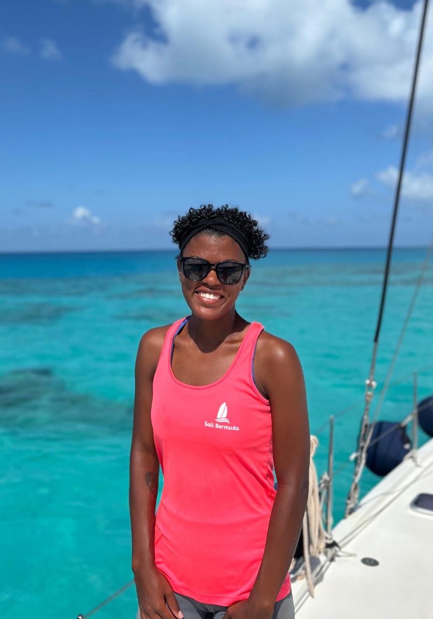 A woman wearing sunglasses and a bright pink tank top stands smiling on a boat, with clear turquoise ocean water and a blue sky in the background.