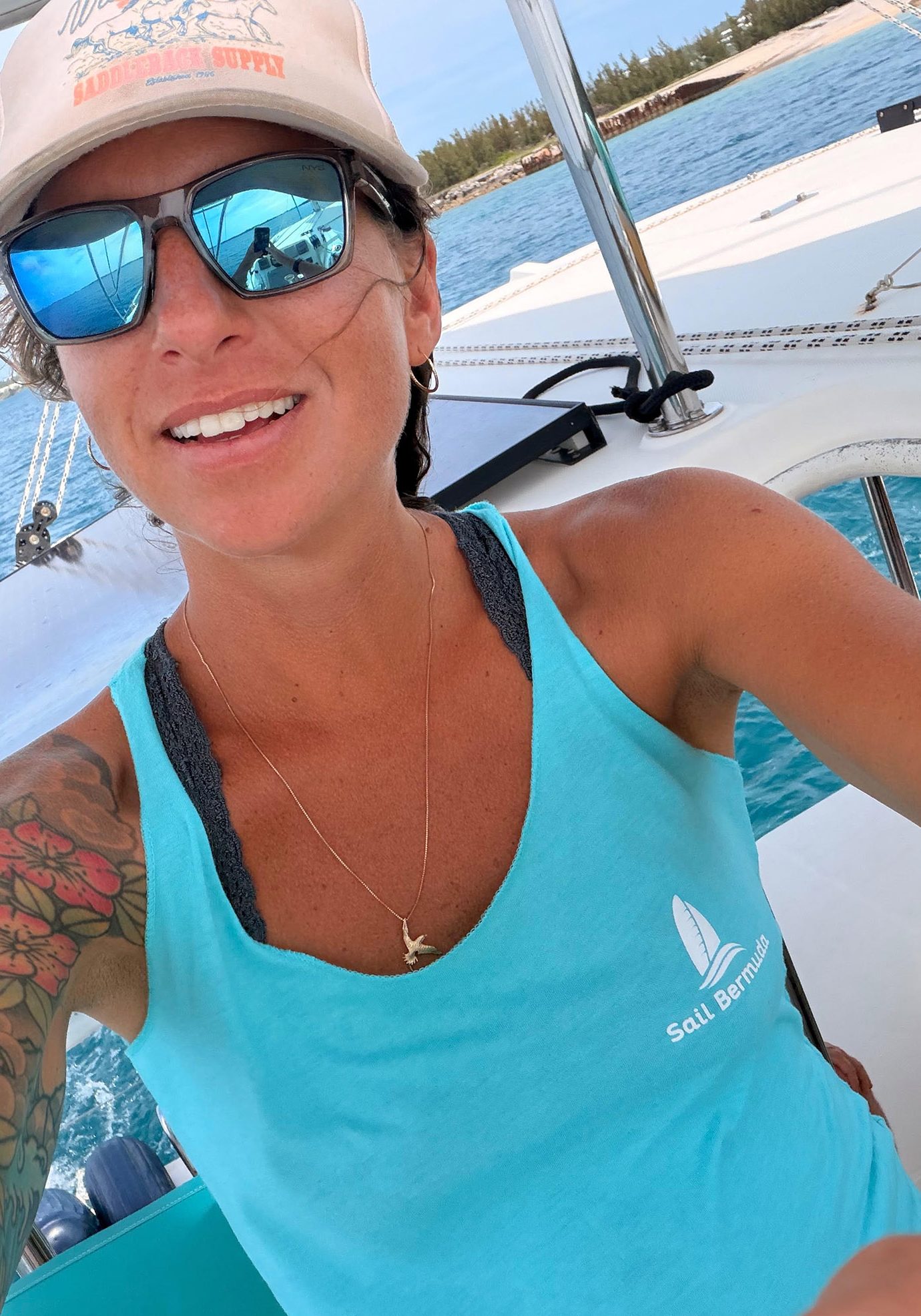 A woman wearing sunglasses, a hat, and a blue Sail Bermuda tank top smiles on a boat, with the ocean and distant shoreline in the background.