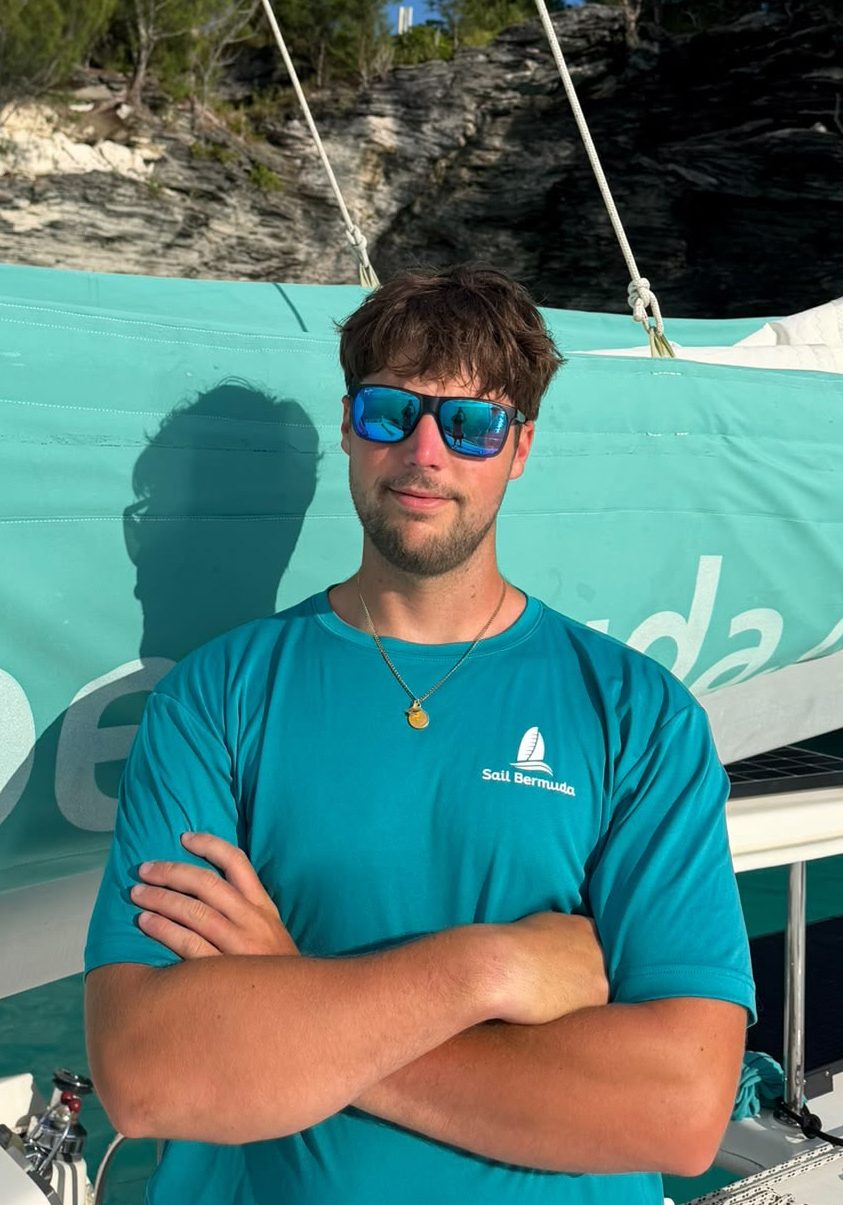 A man in a teal Sail Bermuda shirt and sunglasses stands with arms crossed on a boat, with a teal sail and rocky, forested background in sunlight.