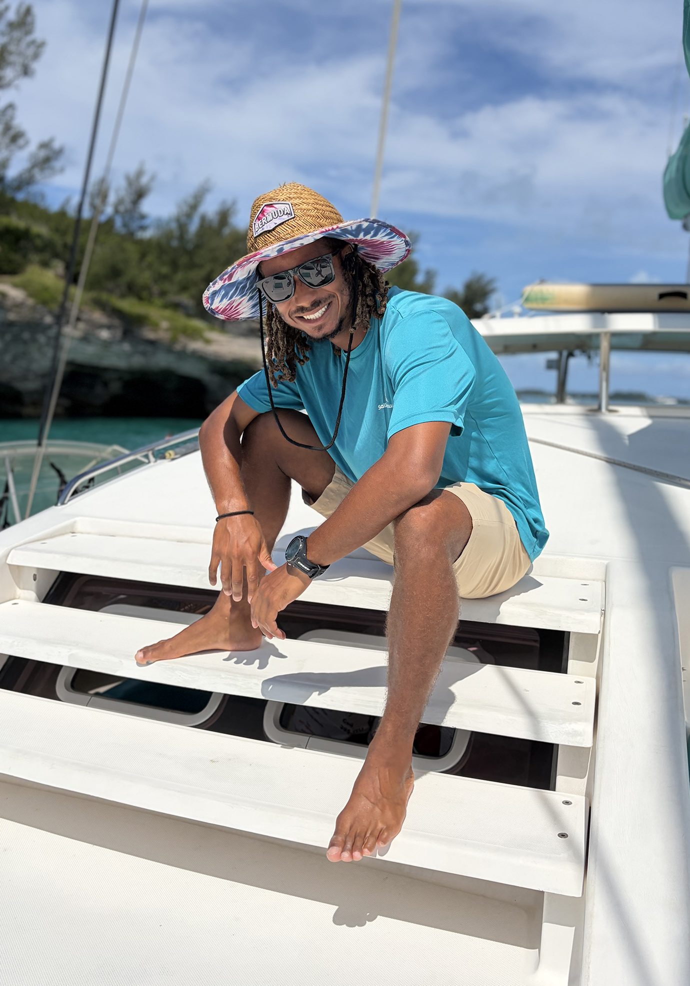 A man in a straw hat and sunglasses sits smiling on the steps of a boat, wearing a turquoise shirt and tan shorts, with blue skies and greenery in the background.