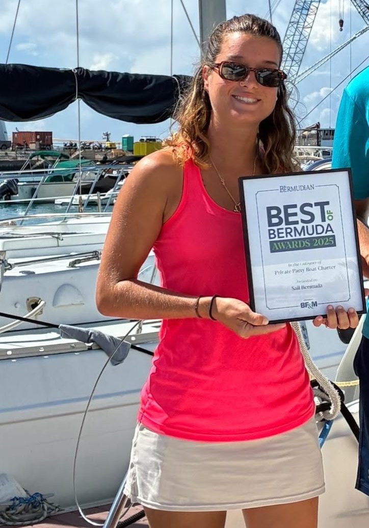 A woman in a pink tank top and sunglasses holds a Best of Bermuda Awards 2025 certificate while smiling in front of docked sailboats on a sunny day.